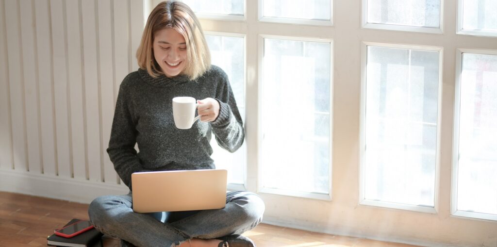Woman sitting on the floor sipping on her drink and working on her laptop